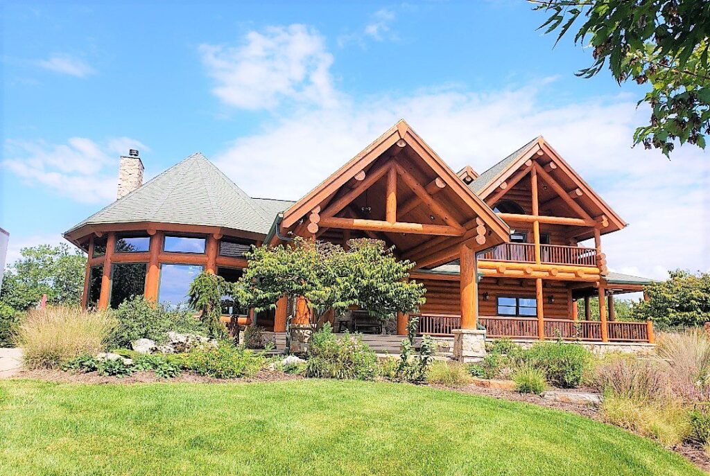 A large log cabin with a unique design, featuring a green roof and expansive porches, surrounded by lush landscaping and blue skies.