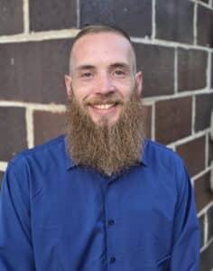 In this image, there is a strawberry blonde pale man with a trimmed beard and mustache in deep blue dress shirt standing in front of a brown brick building. He is smiling.