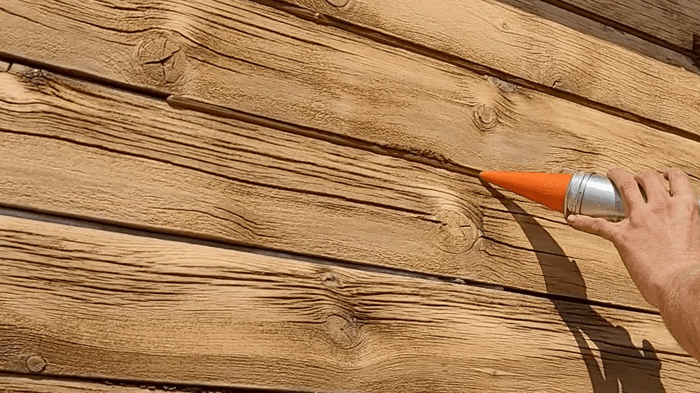 In this image you see someone is caulking log joints on a cabin- you can see a person using a bulk loader to apply.