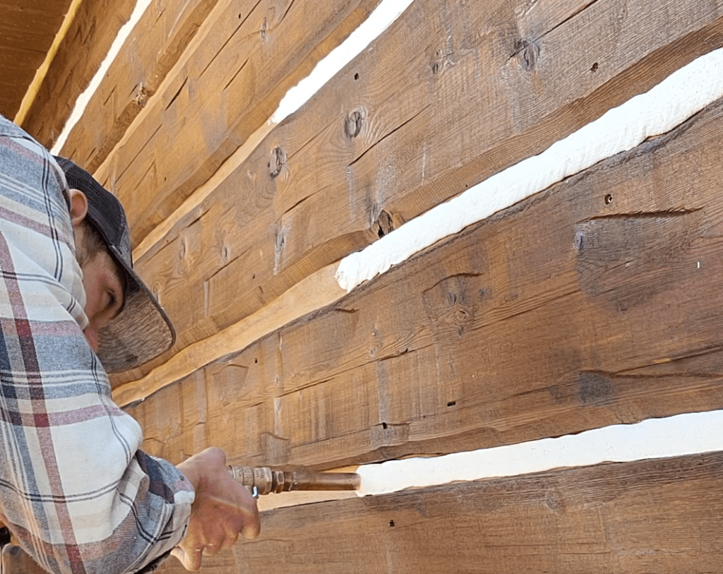 In this image, a Log Master is using a chinking machine to apply a layer of chinking on a log home.