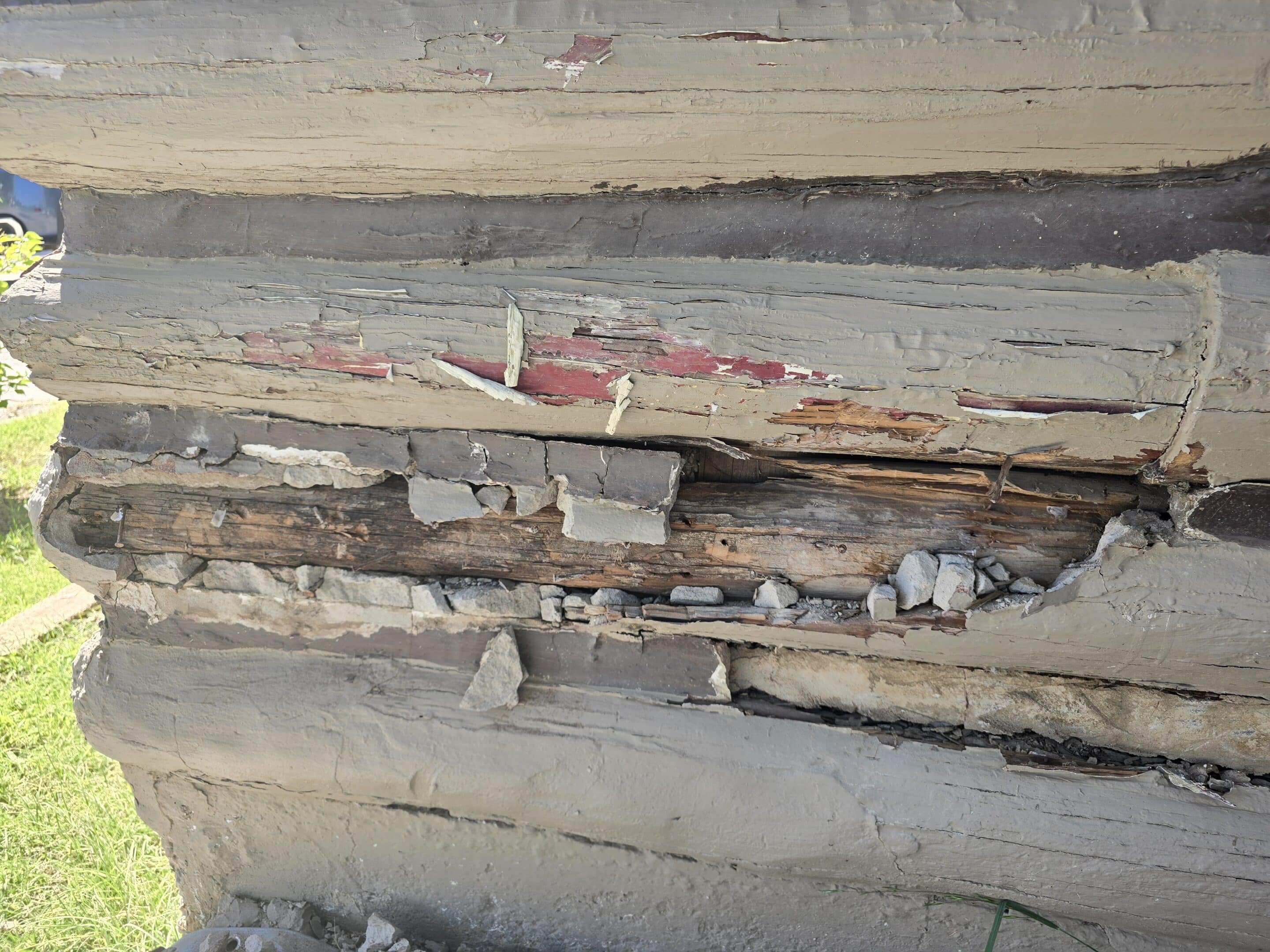 Close-up of a log wall with deteriorated wood, broken mortar chinking, and peeling coatings.